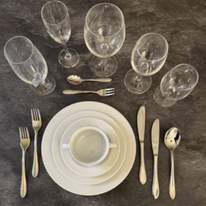 Elegant gedeckter Tisch mit mehreren Gläsern, Besteck und weißem Geschirr, darunter Teller und eine Tasse, auf einem dunklen Untergrund. Ideal für festliche Anlässe oder besondere Dinner-Events.